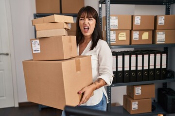 Wall Mural - Young brunette woman working at small business ecommerce holding packages angry and mad screaming frustrated and furious, shouting with anger. rage and aggressive concept.