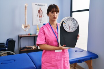 Poster - Young brunette woman as nutritionist holding weighing machine thinking attitude and sober expression looking self confident