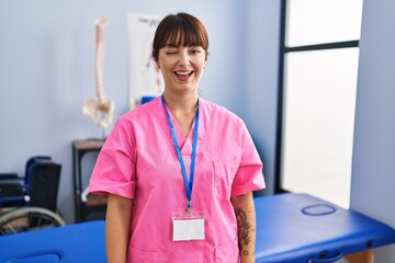 Canvas Print - Young brunette woman working at rehabilitation clinic winking looking at the camera with sexy expression, cheerful and happy face.