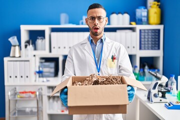 Poster - Young hispanic man working at scientist laboratory holding cardboard box clueless and confused expression. doubt concept.