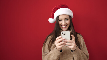 Beautiful hispanic woman wearing christmas hat using smartphone over isolated red background