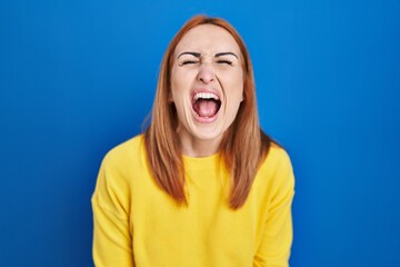 Canvas Print - Young woman standing over blue background angry and mad screaming frustrated and furious, shouting with anger. rage and aggressive concept.