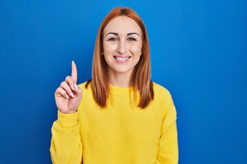 Canvas Print - Young woman standing over blue background showing and pointing up with finger number one while smiling confident and happy.