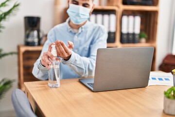 Wall Mural - Young hispanic man business worker wearing medical mask using sanitizer gel hands at office