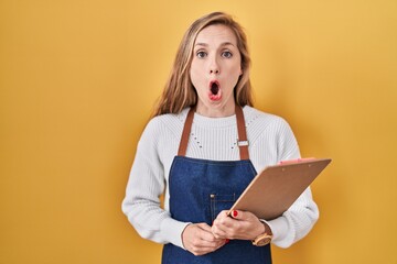 Poster - Young blonde woman wearing professional waitress apron holding clipboard scared and amazed with open mouth for surprise, disbelief face