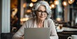 © ibreakstock - Senior adult woman in glasses using laptop, older woman in her 50s with grey hair working on laptop computer in cafe at table