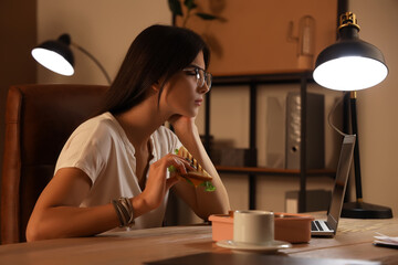 Poster - Young businesswoman with tasty sandwich working in office at night