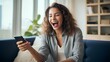 © yj - Excited happy young woman holding smart phone device sitting on sofa at home. Happy satisfied female looking at mobile smartphone screen, gesturing and shout yes. generative AI