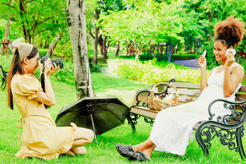 Wall Mural - Asian female friend photographer sitting and taking shot photo, portrait healthy pregnant african american woman sitting on wooden bench in park showing gloves baby smiling looking at camera happy.