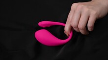 Woman holding a pink vibrating sex toy on a black silk sheet. 