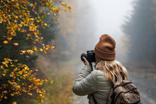 Woman With Camera Taking Picture Of Autumn Leaf. Tourist Hiking In Misty Forest. Landscape Photographer With Backpack Enjoying Nature In Fall Season