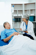 © Nuttapong punna - Friendly Female Head Nurse Making Rounds does Checkup on Patient Resting in Bed. She Checks tablet while Man Fully Recovering after Successful Surgery in hospital