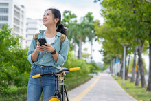 Young beautiful Asian woman is using a smartphone while riding a bicycle through a city park. Lifestyle and environmentally friendly concept.