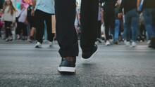 Rear Back View Following Male Feet Steps Crossing Busy Street. Man Wearing Casual Dark Trousers Leather Sneakers Goes On Pedestrian Crossing In Big City. Low Angle People Crowd Legs Walking Sidewalk