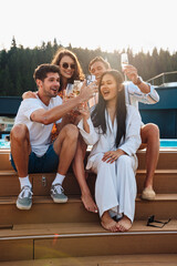 Group of friends drinking champagne and taking selfie while sitting by pool in mountains