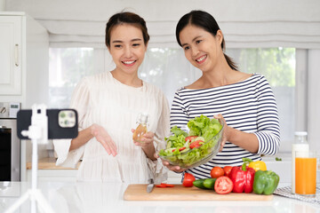 Happy two asian woman friends streaming live video with mobile phone to share online while cooking in kitchen at home