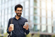© Prostock-studio - Cheerful indian guy going to office, using phone, drinking coffee