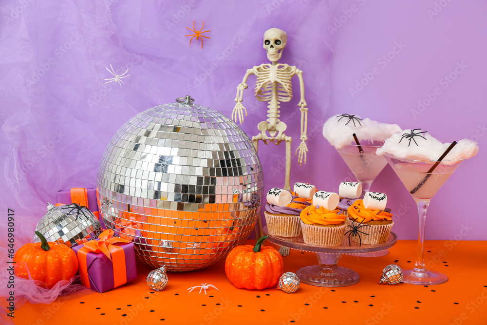 Halloween party composition with cupcakes, disco ball, drinks and decorations on orange table against purple background