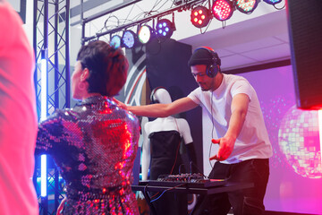 Young man dj mixing techno tracks on stage while people partying on dancefloor in nightclub. Musician playing electronic music on stage while clubbers dancing at discotheque