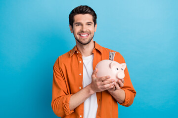 Poster - Portrait of cheerful wealthy nice man toothy smile hands hold money bank pig dollar bills isolated on blue color background