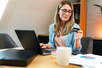 Wall Mural - Blonde woman with eyeglasses sitting at a table in a dining room and online shopping from home on her laptop