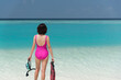 Back of Asian woman in pink swimsuit standing with mask and fins enjoy looking view of beautiful sea on hot summer day. Beautiful fit body girl in swimsuit relax on travel. Summer vacation concept.