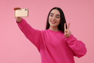 Canvas Print - Smiling young woman taking selfie with smartphone and showing piece sign on pink background, space for text