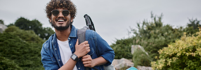 happy indian man in sunglasses standing with backpack on green hill, hiker with travel gear, banner