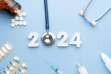 2024 Happy New Year banner for health care and medical concept. Stethoscope with doctor pills, syringe, vaccine vial, eyes drop, antibiotics and white number 2024 on table blue background.