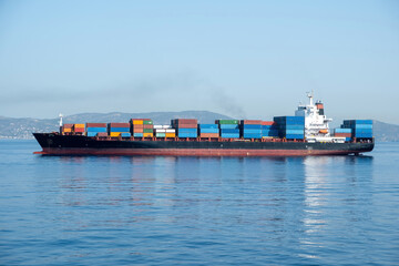 Wall Mural - Container cargo ship loaded leaves Piraeus port Greece. Storage and carry. Reflection in blue sea