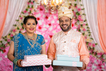 Portrait shot of happy smiling Middle aged couple with Gift boaxes in hand posing to to camera on wedding stage - concept of marriage moment, Traditional ethnic wear and Special occasion.