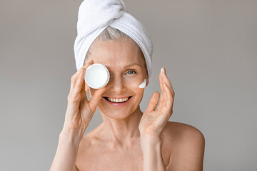 Beautiful mature woman holding jar with moisturising cream in hand, covering one eye with skincare product