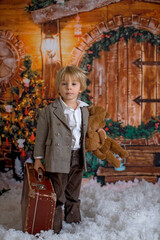 Poster - Cute fashion toddler boy, playing in the snow with teddy bear in front of a wooden cabin log