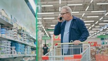 An elderly man walking through a supermarket with a trolley and talking on the phone. Pensioner in a grocery shop