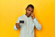 African American man with credit card, yellow studio, pointing temple with finger, thinking, focused on a task.