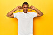 © Asier - African American man in white polo, yellow studio, covering ears with fingers, stressed and desperate by a loudly ambient.