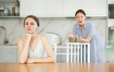 Upset daughter is sitting at table. Mom yells at her daughter in the background