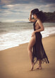 Captivating female model with curly hair and a slender sexual body poses elegantly in a chic black beach dress and hat, showcasing her attractive physique at the tropical beach.