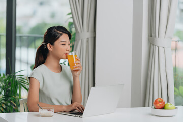 Wall Mural - Beautiful young woman eating oatmeal and drinking orange juice while working with laptop