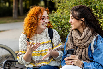 two woman friends sitting on bench in public park with mobile phone on hand, talk and smile