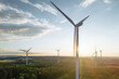 Wind turbines in a hilly forest in front of a partly cloudy, but sunny sky are seen from an aerial view during sunset