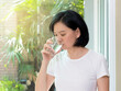 Beautiful Asian woman with clean skin, natural make-up, short black hair drinking water at home. Middle aged woman holding transparent glass in her hand. Image for skin and healthy concept, copy space