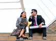 Happy Asian business man and woman sitting on the first step talking about the future. Young co-workers on breaking time relaxing outdoor with the background of urban landscape.