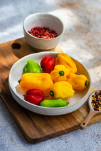 Close-up Of A Bowl Of Red, Yellow And Green Scotch Bonnet Peppers On A Chopping Board With Chilli Flakes And Pink Peppercorns