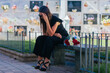 Weeping woman with sunglasses, black dress and hand on her head, sitting on a bench, with a bouquet of roses beside her, in a cemetery.