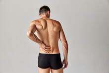 Back Pains. Muscular Man Standing In Underwear And Holding His Back Against Grey Studio Background. Medical Treatment. Concept Of Men's Health And Beauty, Body Care, Fitness, Wellness, Ad