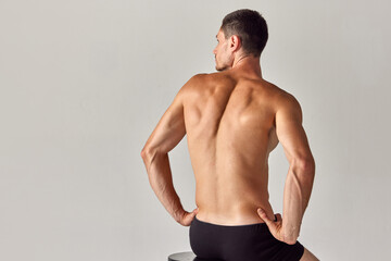 Back view image of muscular young shirtless man with relief, sign back sitting in underwear against grey studio background. Concept of men's health and beauty, body care, fitness, wellness, ad