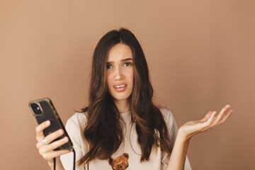 Wall Mural - Mad and shock young brunette beautiful woman using mobile phone isolated on beige background. Yelling unhappy woman hold mobile phone. Disappointed sad upset lady horrified impressed news. WTF. Oh no.