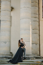 Newlyweds Walk Near Baroque Roman Catholic Church In Pidhirtsi Ukraine. Bride And Groom Kissing On Stairs Near Large Columns Of Ancient Temple At Sunset. Couple Hold Hands Nature Autumn Day. Back View
