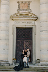 Parents hugging kids near at Catholic church of Saint Joseph in Pidhirtsi. Mother, father, daughter, son stands on steps near large columns of ancient temple at sunset. Family spending time together